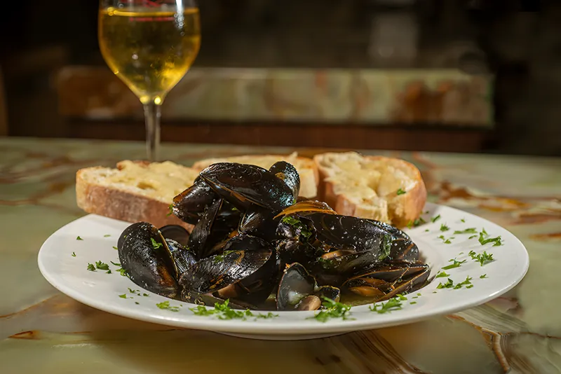A plate of mussels in white wine and garlic with toasted pizza crostini, a glass of white wine behind